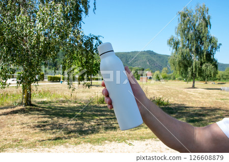 Man holding in the hand white thermo stainless bottle with refreshing water on the nature. 126608879