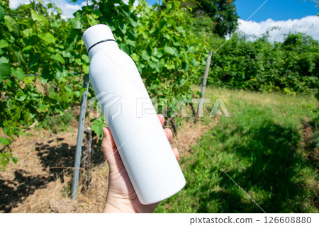 Man holding in the hand white thermo stainless bottle with refreshing water on the nature. 126608880
