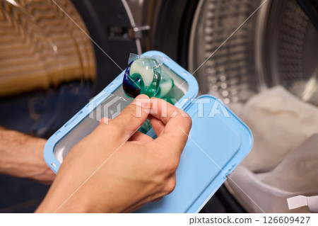 A hand is holding a colorful laundry pod as it is placed into a washing machine for cleaning A hand is holding a colorful laundry pod as it is placed into a washing machine for cleaning 126609427