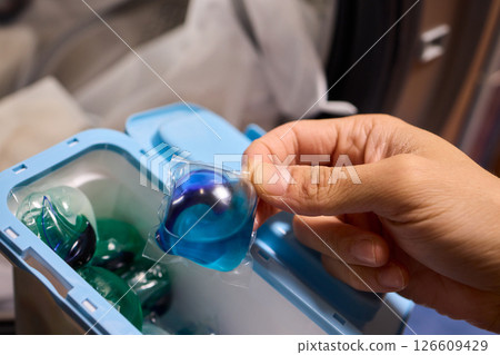 A hand is holding a colorful laundry pod as it is placed into a washing machine for cleaning 126609429