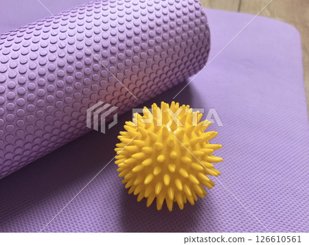 Sports equipment shown close-up: yellow massage ball and purple myofascial release roller behind it against the background of yoga, pilates and fitness mat lying on wooden floor. Fitness and health 126610561