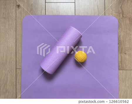 Top view of sports equipment: yellow massage ball, purple myofascial release roller and fitness yoga mat lying on wooden floor. Fitness and healthy lifestyle equipment Top view of sports equipment: yellow massage ball, purple myofascial release roller and fitness yoga mat lying on wooden floor. Fitness and healthy lifestyle equipment 126610562