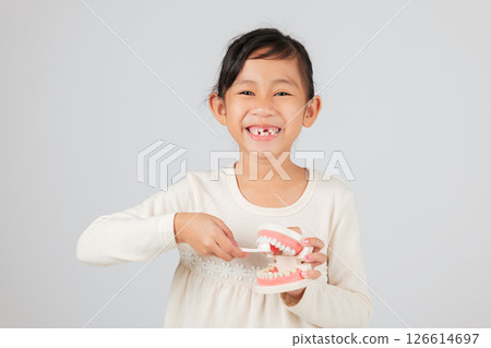 Dentist teaches Asian kid about tooth loss, holding toothbrush and teeth model. Girl smiles with gap, showing healthy dental habits and missing milk teeth. isolated white background Dentist teaches Asian kid about tooth loss, holding toothbrush and teeth model. Girl smiles with gap, showing healthy dental habits and missing milk teeth. isolated white background 126614697