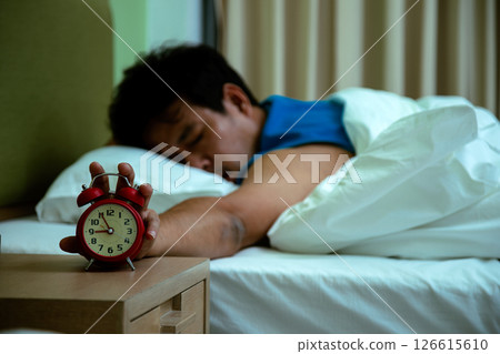 Sleeping man in bed reaches for ringing alarm clock, representing early morning wake-up or reluctance to get up Sleeping man in bed reaches for ringing alarm clock, representing early morning wake-up or reluctance to get up 126615610