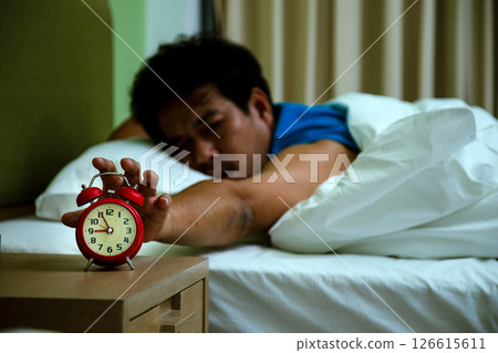 Sleepy man in bed stretching to turn off a red alarm clock on bedside table, illustrating morning wake-up struggle Sleepy man in bed stretching to turn off a red alarm clock on bedside table, illustrating morning wake-up struggle 126615611