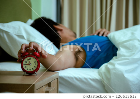Sleepy man lying in bed reaching out to turn off a ringing red alarm clock, representing morning tiredness and reluctance to wake up 126615612