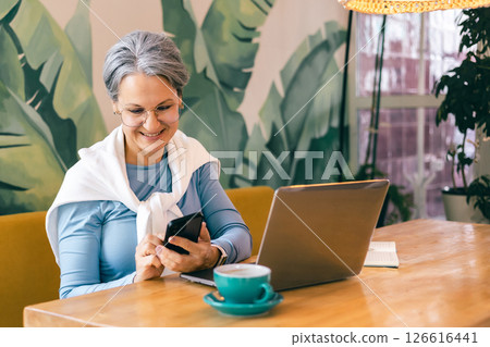 Happy senior freelancer woman using smartphone while working from cozy cafe with laptop 126616441