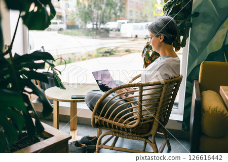 Woman with laptop sitting in cozy cafe near large window with greenery, working on a sunny day 126616442