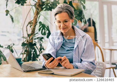 Senior freelance woman using smartphone and writing notes while working remotely in a cozy cafe Senior freelance woman using smartphone and writing notes while working remotely in a cozy cafe 126616465
