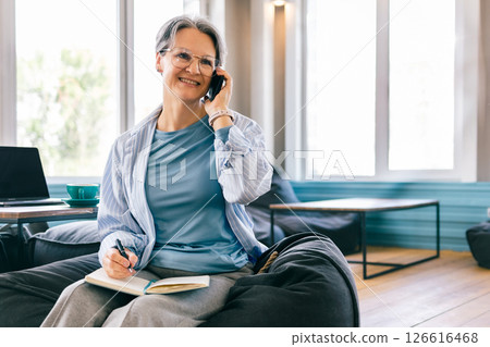 Happy senior woman on phone call while journaling, balanced lifestyle and wellness during menopause 126616468