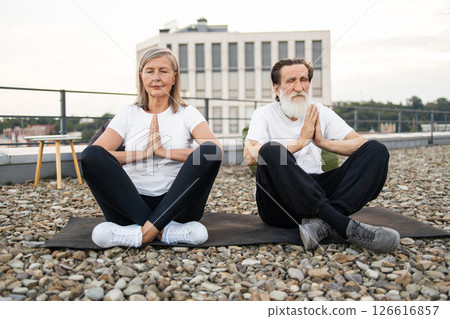 Senior man and woman performing seated yoga meditation outdoors on rooftop setting. Engaged in mindful namaste pose in serene atmosphere reflecting fitness, relaxation, and harmony. 126616857