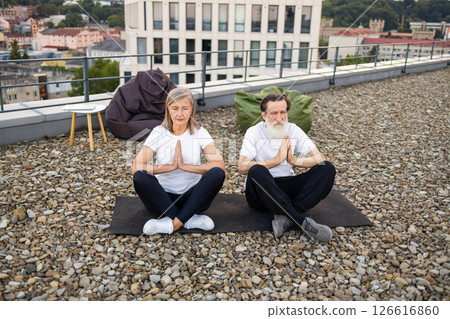 Senior man and woman practicing yoga namaste pose on yoga mats outdoors on rooftop. Senior man and woman practicing yoga namaste pose on yoga mats outdoors on rooftop. 126616860