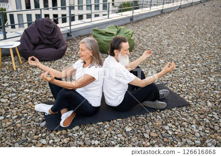 Senior couple performing relaxing meditation seated back to back in lotus pose outdoors. Man and woman wearing white and black activewear. Balanced lifestyle and harmony concept in urban environment. 126616868