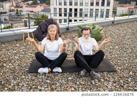 Senior man and woman in white tees practicing yoga meditation in lotus pose outdoors on rooftop under clear sky. Prominent relaxed and peaceful expressions. 126616876