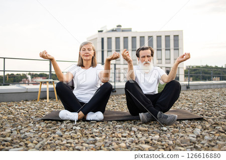 Senior man and woman practicing meditation together, sitting in lotus pose on rooftop stones. Emitting calmness, peaceful energy. Comfortable clothing fits outdoor artistic environment urban buildings Senior man and woman practicing meditation together, sitting in lotus pose on rooftop stones. Emitting calmness, peaceful energy. Comfortable clothing fits outdoor artistic environment urban buildings 126616880