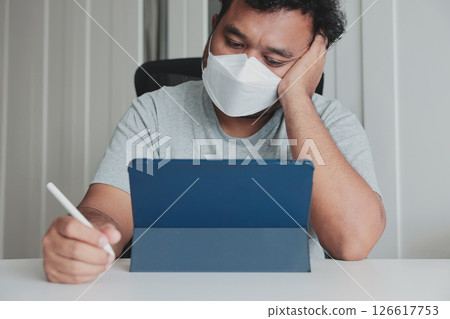 Bored man wearing face mask working from home with tablet and stylus during pandemic, tired and unmotivated, remote work burnout, lack of focus, pandemic fatigue, digital lifestyle. 126617753