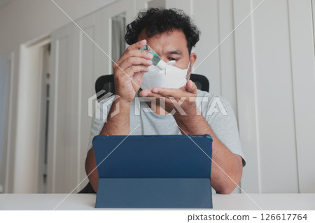Man in face mask sanitizing hands while working from home, using alcohol gel for hygiene and protection during the pandemic in modern indoor setting. 126617764