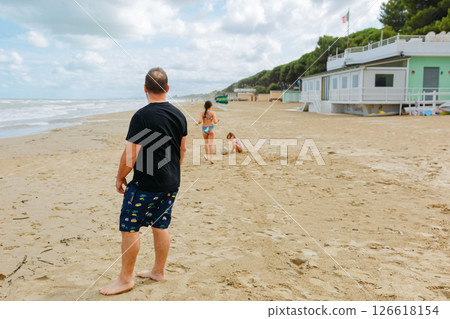 rear view of a man standing on a sandy beach and watching at his children playing in front 126618154