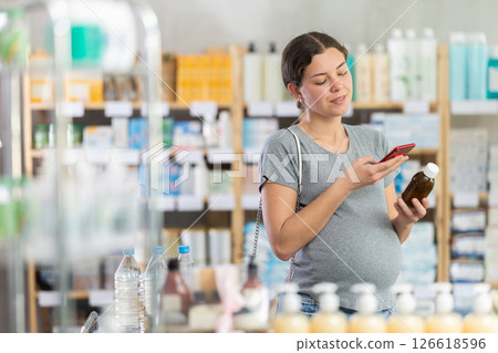 Young pregnant female buyer choosing medicinal cough liquid syrup standing between shelves at pharmacy, scan QR code on label 126618596