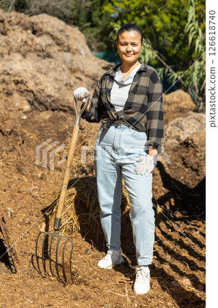 Asian girl stands against background of large pile of manure, leans on pitchfork 126618740