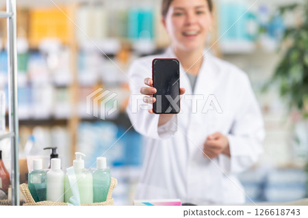 Friendly skilled young girl pharmacist standing behind counter, demonstrating and offering mobile application for smartphone in pharmacy Friendly skilled young girl pharmacist standing behind counter, demonstrating and offering mobile application for smartphone in pharmacy 126618743