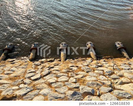 Five drainage pipes lined along a rocky embankment, discharging into a lake under the golden sunlight, showcasing water management infrastructure and engineering Five drainage pipes lined along a rocky embankment, discharging into a lake under the golden sunlight, showcasing water management infrastructure and engineering 126618830