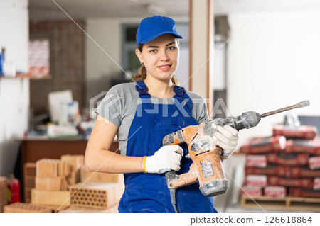 Female builder in blue overalls holding pneumatic jackhammer at construction site Female builder in blue overalls holding pneumatic jackhammer at construction site 126618864