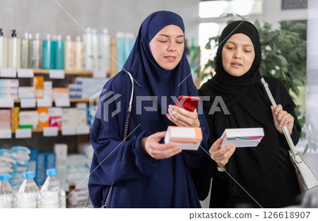 Attentive two woman in hijab choosing box with pills to improve well-being in pharmacy, scan QR code on label Attentive two woman in hijab choosing box with pills to improve well-being in pharmacy, scan QR code on label 126618907