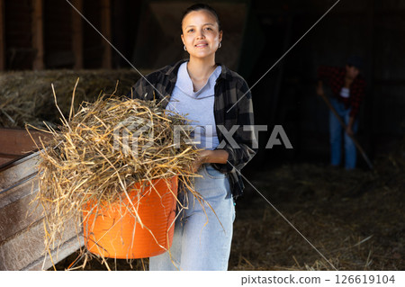 Asian young female worker walks go through corridor of stable, carrying basket with hay Asian young female worker walks go through corridor of stable, carrying basket with hay 126619104