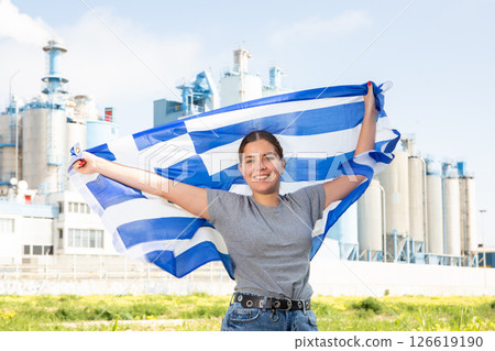 Young female in casual wear waving state flag of Greece while standing in front of big tanks at chemical plant 126619190