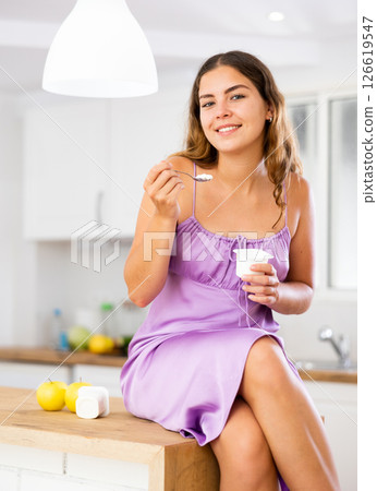 Portrait of positive girl in nightdress eating yogurt at home Portrait of positive girl in nightdress eating yogurt at home 126619547