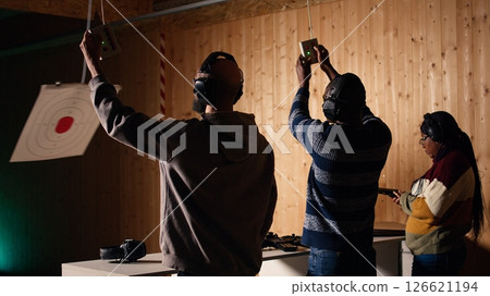 Group of friends in firing range doing recreational shooting activity with firearm, hitting bullseyes. People in shooting gallery training with handguns, sharpening skills, camera A 126621194