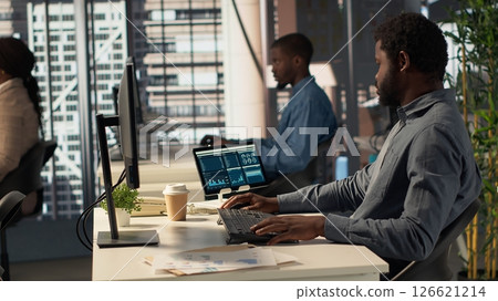 Black analyst working with monthly revenue balance to create financial plans using online tools, analyzing data infographics. Businessman managing organizational goals for efficiency. Camera A. 126621214