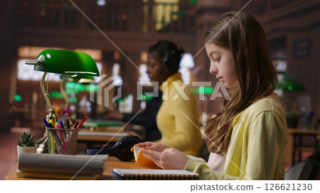 Focused schoolgirl reading a book in the library for mandatory literature, gaining knowledge for her academic curriculum during study time. Young scholar focusing on lecture tasks. Camera B. 126621230