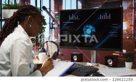 Remote worker at home watching business conference on PC monitor, looking over statistics on documents. Freelancer in apartment looking at presentation and checking paperwork files, camera A 126621289