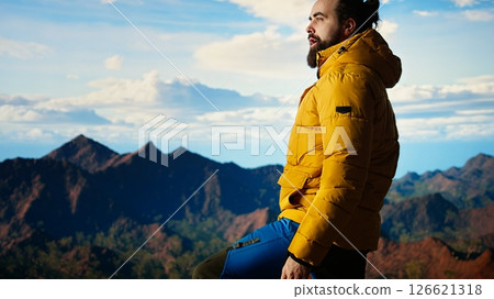 Adventure seeker gazes at the valley and mountain peaks ahead, feeling the freedom of the open wilderness. Young man fully equipped enjoying high altitude outdoor exploration. Camera A. 126621318