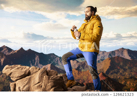 Young man explorer climbing the mountain range and hydrating with his water bottle, drinking refreshment on top of the rocky mountain. Male hiker refreshing after the trekking activity. Young man explorer climbing the mountain range and hydrating with his water bottle, drinking refreshment on top of the rocky mountain. Male hiker refreshing after the trekking activity. 126621323