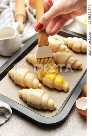 Baker brushing egg yolk on raw croissants on baking sheet 126621378
