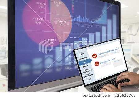 Someone working on notebook while a screen in the background showing graph of working results. 126622762