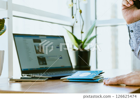 Dubai, UAE- 07.05.2025: A man sits at a table and plans a vacation with his family through the website booking.com, a man holds Kazakhstan passports and books a hotel through a computer 126625474