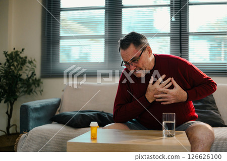 Elderly male experiencing discomfort, hand on chest with medication bottle on the table Elderly male experiencing discomfort, hand on chest with medication bottle on the table 126626100