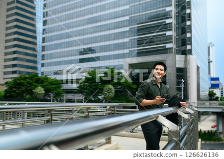 Smiling businessman leaning on railing in an urban area with smartphone in hand Smiling businessman leaning on railing in an urban area with smartphone in hand 126626136