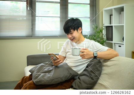 Asian male smiling while checking smartphone and holding a coffee mug on sofa Asian male smiling while checking smartphone and holding a coffee mug on sofa 126626162