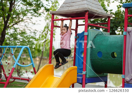 happy toddler girl hanging the red bar by her hand at outdoor playground in park 126626330