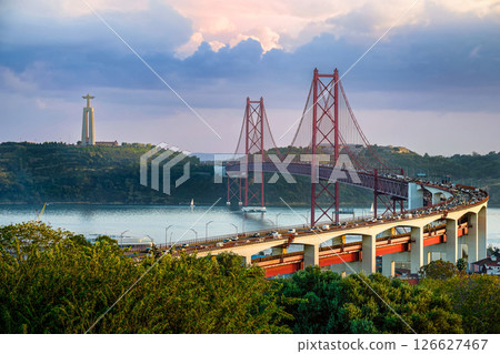 Scenic view of Lisbon iconic 25 de Abril Bridge over Tagus River with Cristo Rei or Christ the King statue in background at sunset. City traffic, lush greenery, tranquil river, famous landmarks, red 126627467