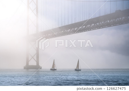 Dramatic view of Lisbon iconic 25 de Abril bridge in morning fog on sunny day, two sailboats navigate calm Tagus river, Portugal. Misty atmosphere, peaceful, quiet scene, mist roll in over waters 126627475
