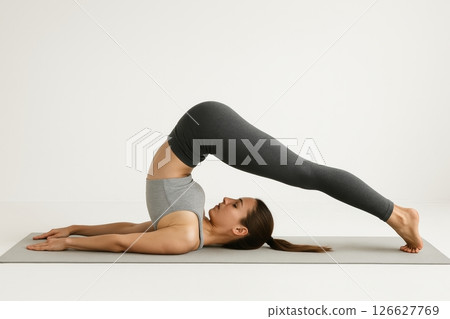 Young caucasian woman practicing plow pose yoga on a mat in a minimalist setting. concept of flexibility, mindfulness, healthy lifestyle, indoor exercise, balance. white background 126627769