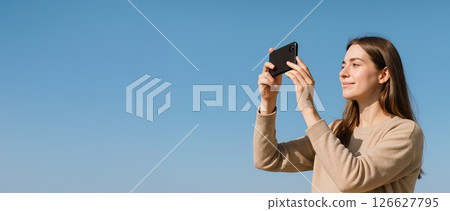 Young caucasian woman photographing outdoors with smartphone against clear blue sky. concept of outdoor photography, technology, travel, creative expression, Copy space, Wide banner 126627795