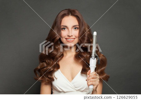 Good-looking young model woman with long curly wavy hairstyle showing hot curling tongs iron Good-looking young model woman with long curly wavy hairstyle showing hot curling tongs iron 126629055