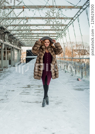 Glamorous brunette woman standing on snow city street wearing hat and fur coat 126629300